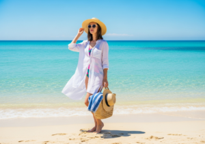 Mladá žena na pláži u tyrkysové vody, v klobouku a brýlích, drží tašku s ručníkem, bosá v písku.